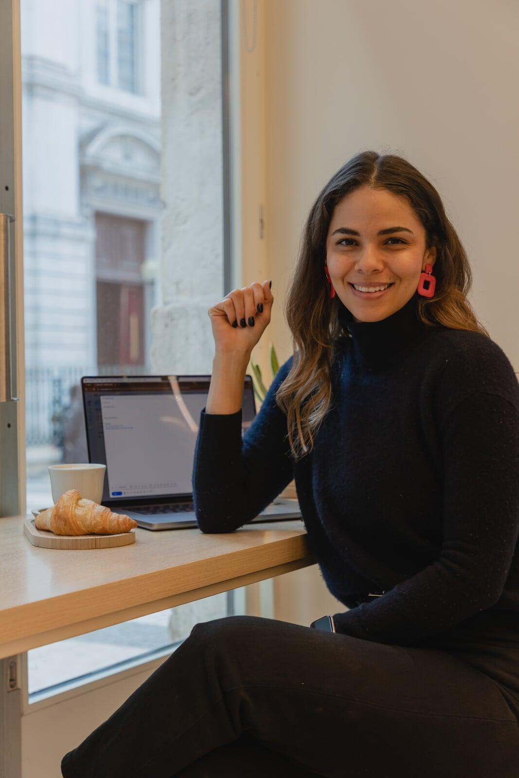 Image of a woman smiling, wearing a black turtle neck, sat next to a coffee and computer in a cafe